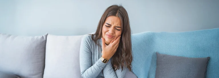 Woman on a couch holding her jaw from sudden tooth pain, a common sign of a dental emergency
